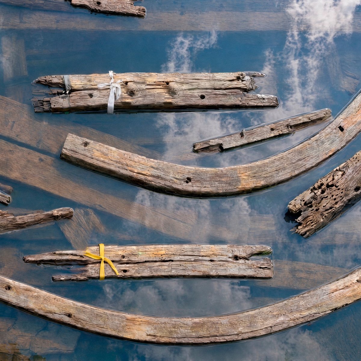 Detail of Riaffioramenti / Resurfacing 01: timber fragments, a yellow ribbon and reflections of clouds in the water